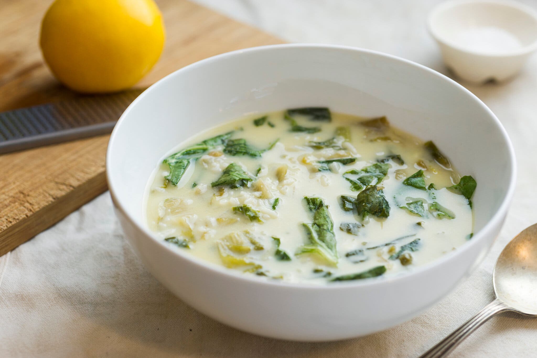 A creamy bowl of Greek rice and escarole soup sits in a white bowl, a lemon and Microplane peaking out in the corner of the frame.