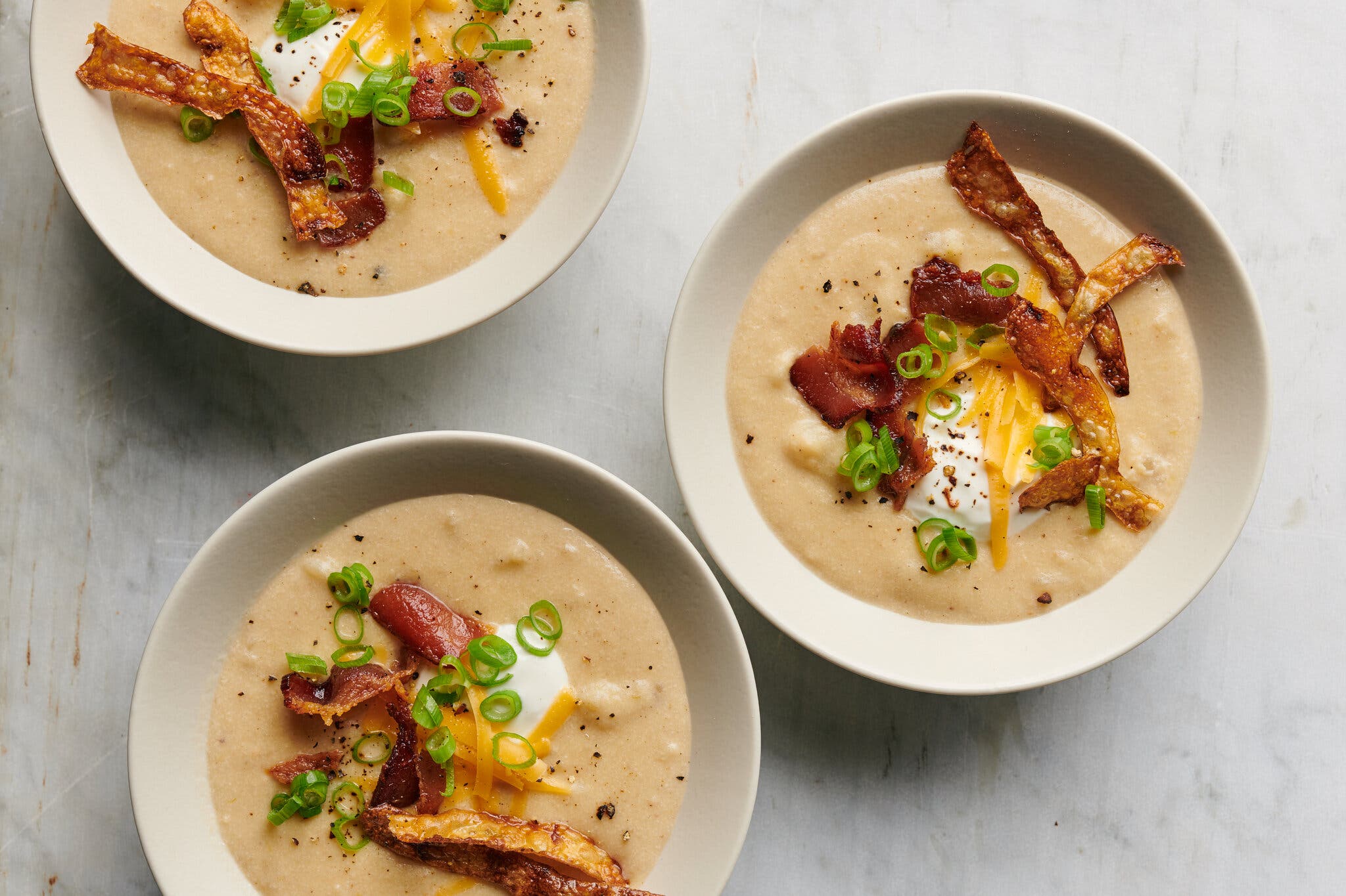 Three bowls of baked potato soup top with cheese, bacon, scallions, shredded cheese and fried potato skins sit on a surface.