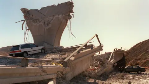 Getty Images A highway has been turned into rubble after an earthquake, with an overpass split in half and two cars abandoned in the rubble