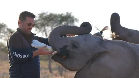 Colossal Biosciences Ben Lam in feeding a baby elephant from a bottle with the Botswanan jungle in the background