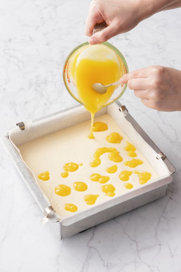 Dropping dollops of bright yellow lemon curd on top of the cheesecake batter.