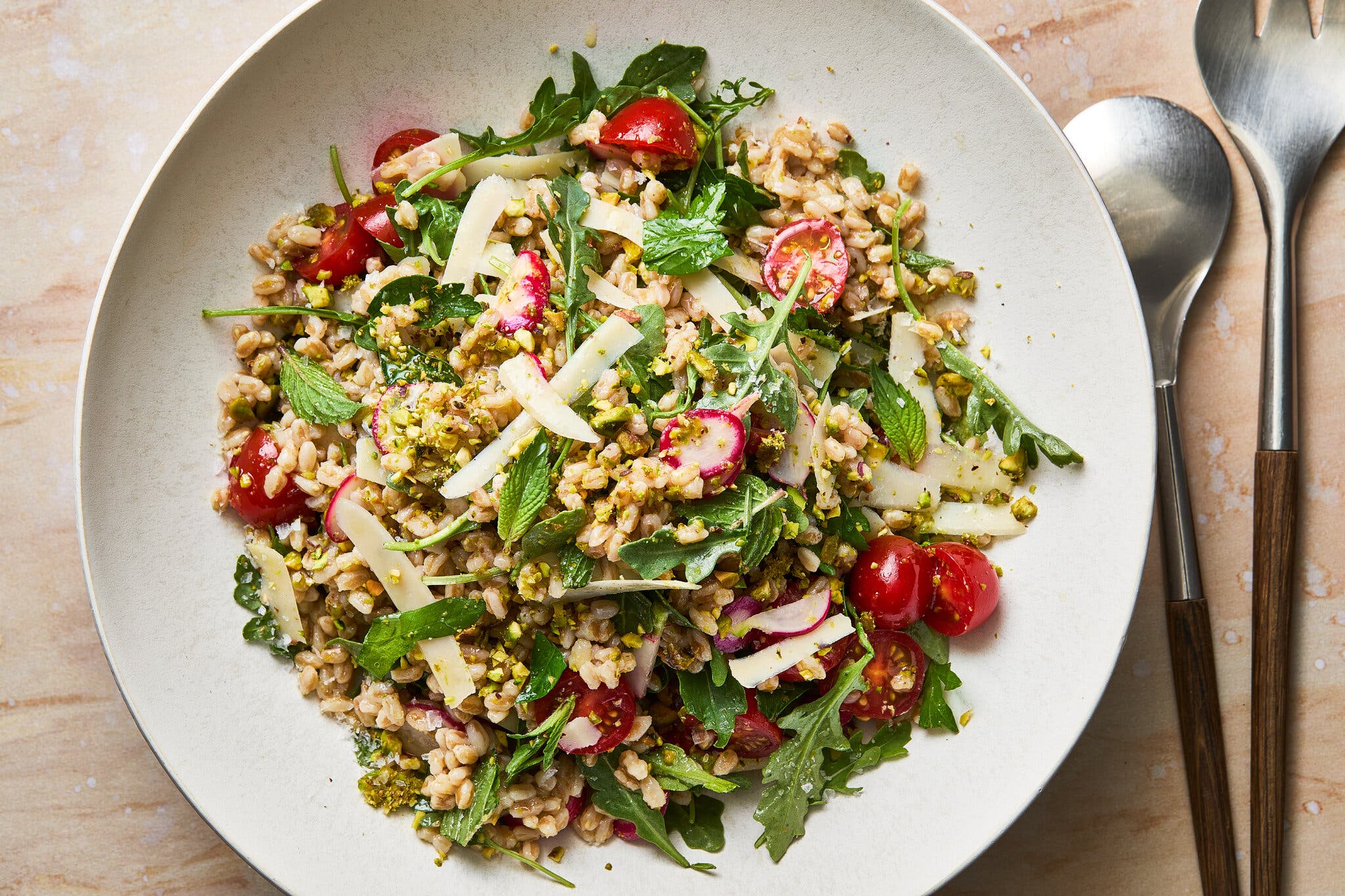 An overhead shot of a farro salad with arugula, cherry tomatoes, and sliced radishes, topped with cheese shavings and pistachios.