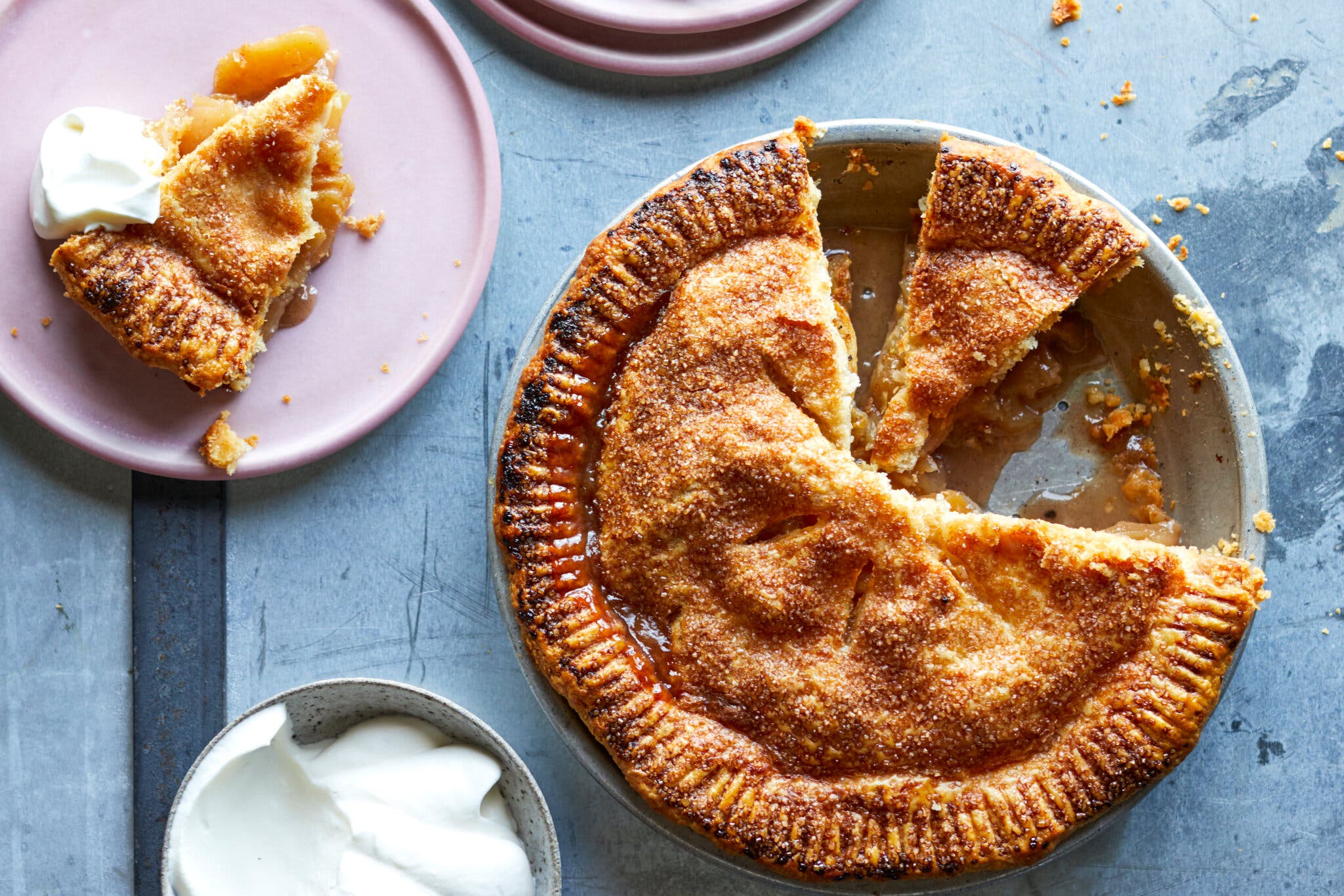 An overhead shot of a golden-brown apple pie with two slices cut out, with one of the slices on a separate plate with whipped cream.