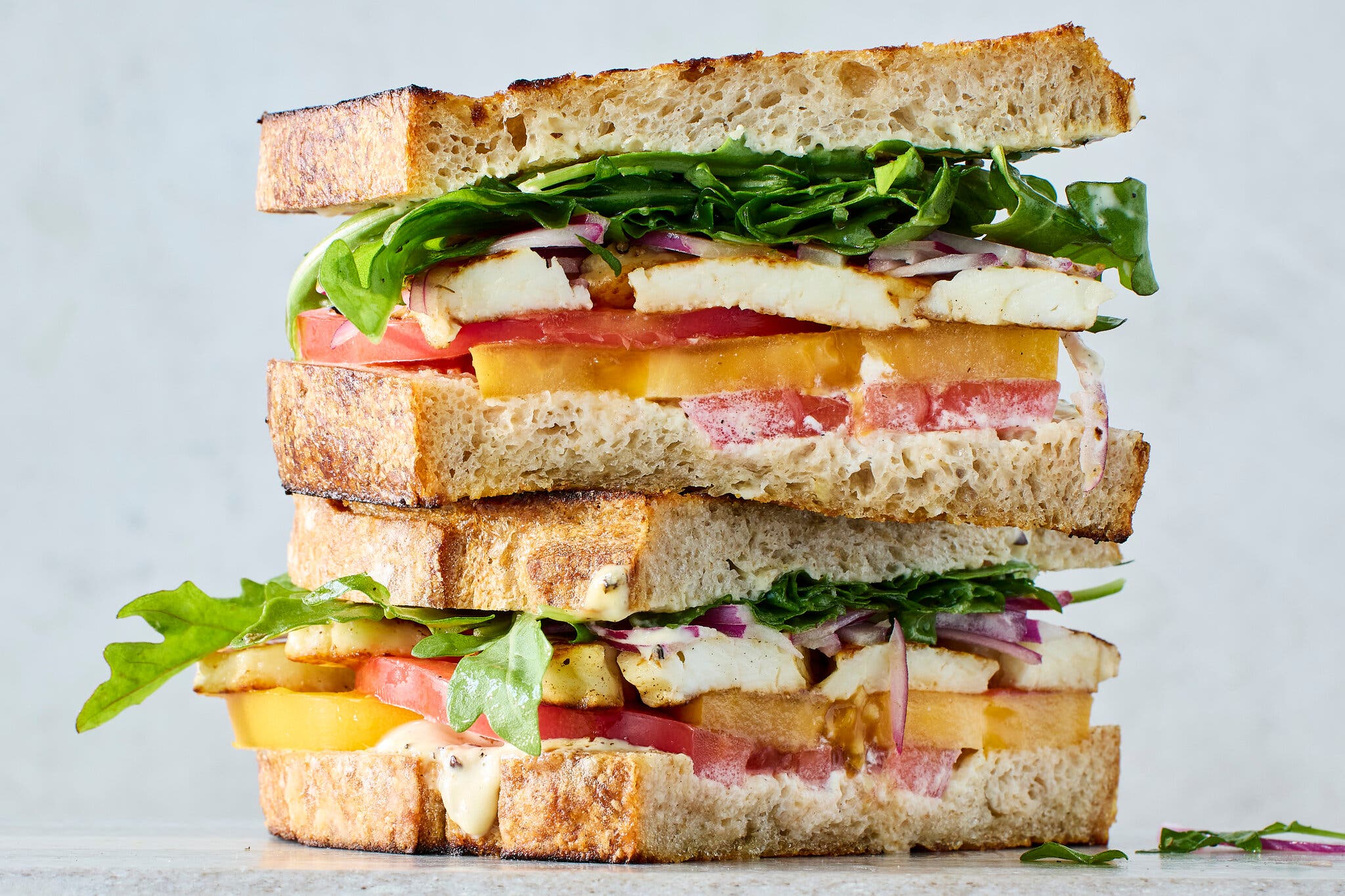 A very tall sandwich, cut in half and stacked with grilled halloumi cheese, sliced yellow and red heirloom tomatoes, and arugula on rustic bread.