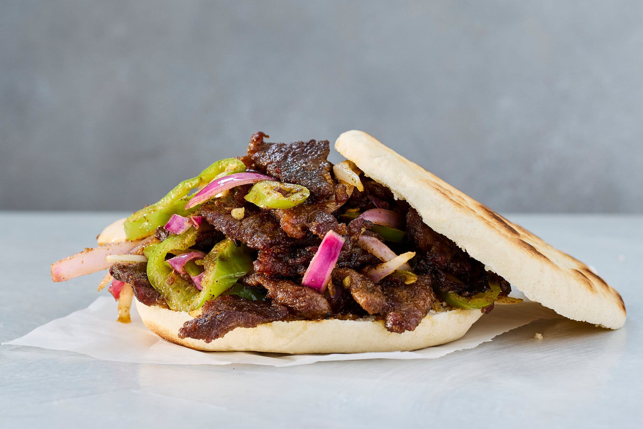 A pita sandwich, with the top half leaning against the filling, overflowing with thinly sliced lamb, sautéed green peppers and red onions.