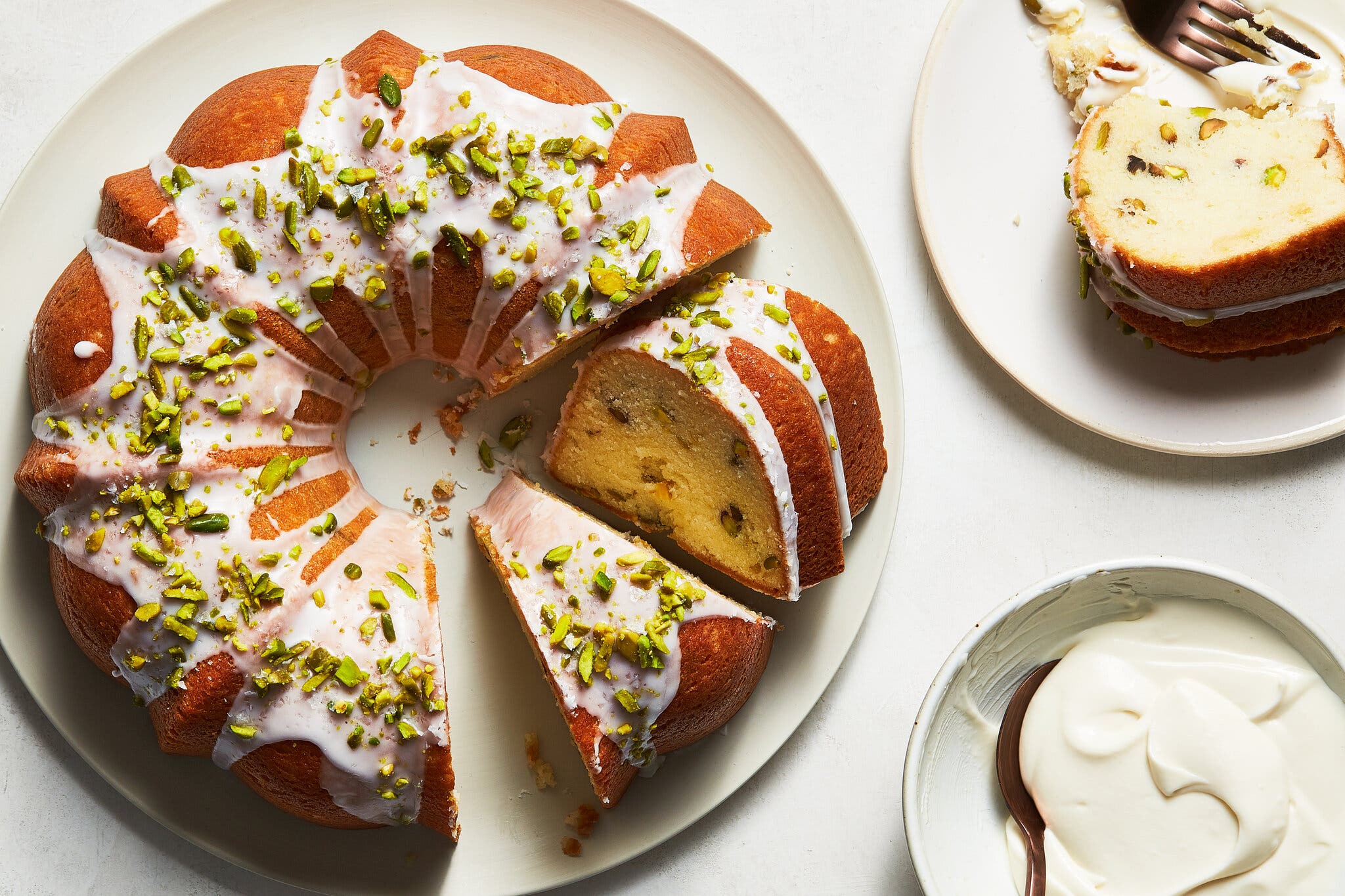 An overhead shot of a pistachio Bundt cake, sliced and drizzled with a white glaze and sprinkled with chopped green pistachios, with a small dish of cream on the side.