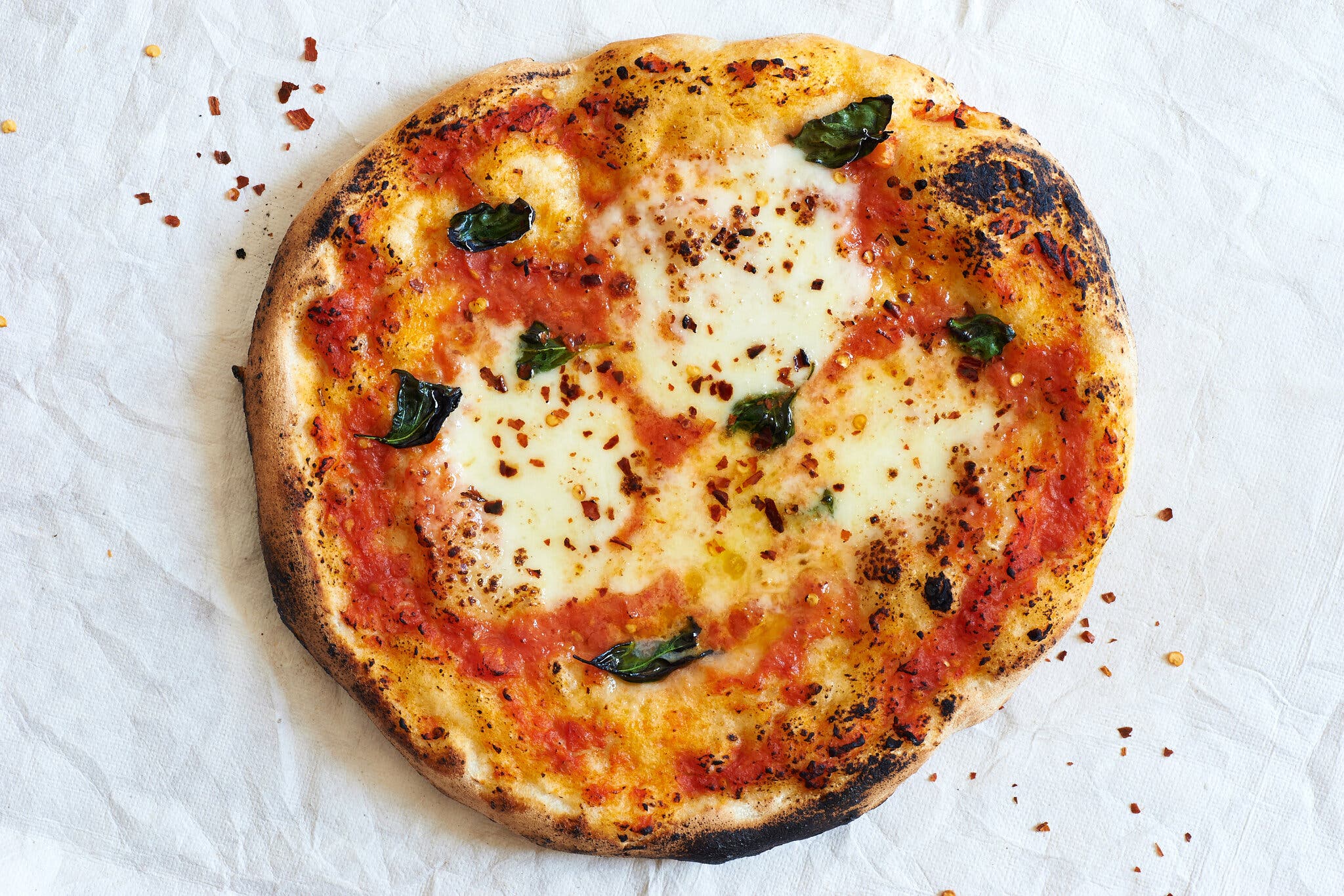 An overhead shot of a round pizza with a charred crust, red sauce, cheese and basil, on a crumpled white background.