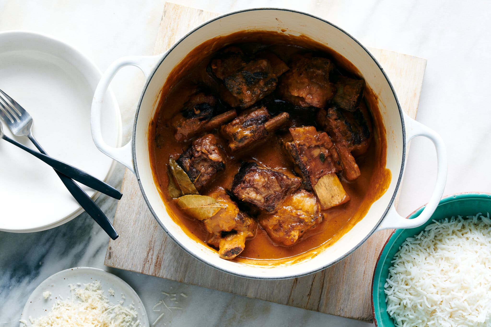 An overhead shot of a large white pot filled with short ribs in a rich brown sauce, with plates and rice on the side.