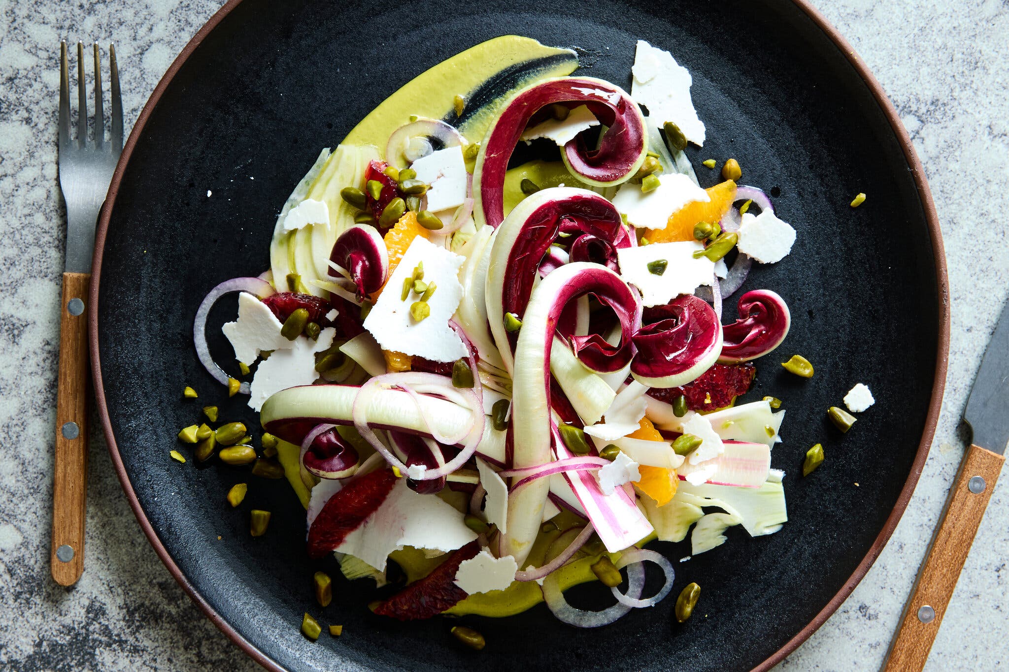 An overhead shot of a radicchio salad with orange slices, nuts and shaved cheese, arranged on a dark plate with a green sauce.