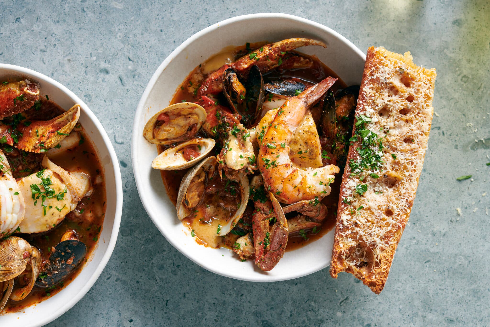 An overhead shot of a large bowl of seafood stew with crab, shrimp, clams and mussels in a reddish-brown broth, with a piece of toasted bread with cheese and herbs.