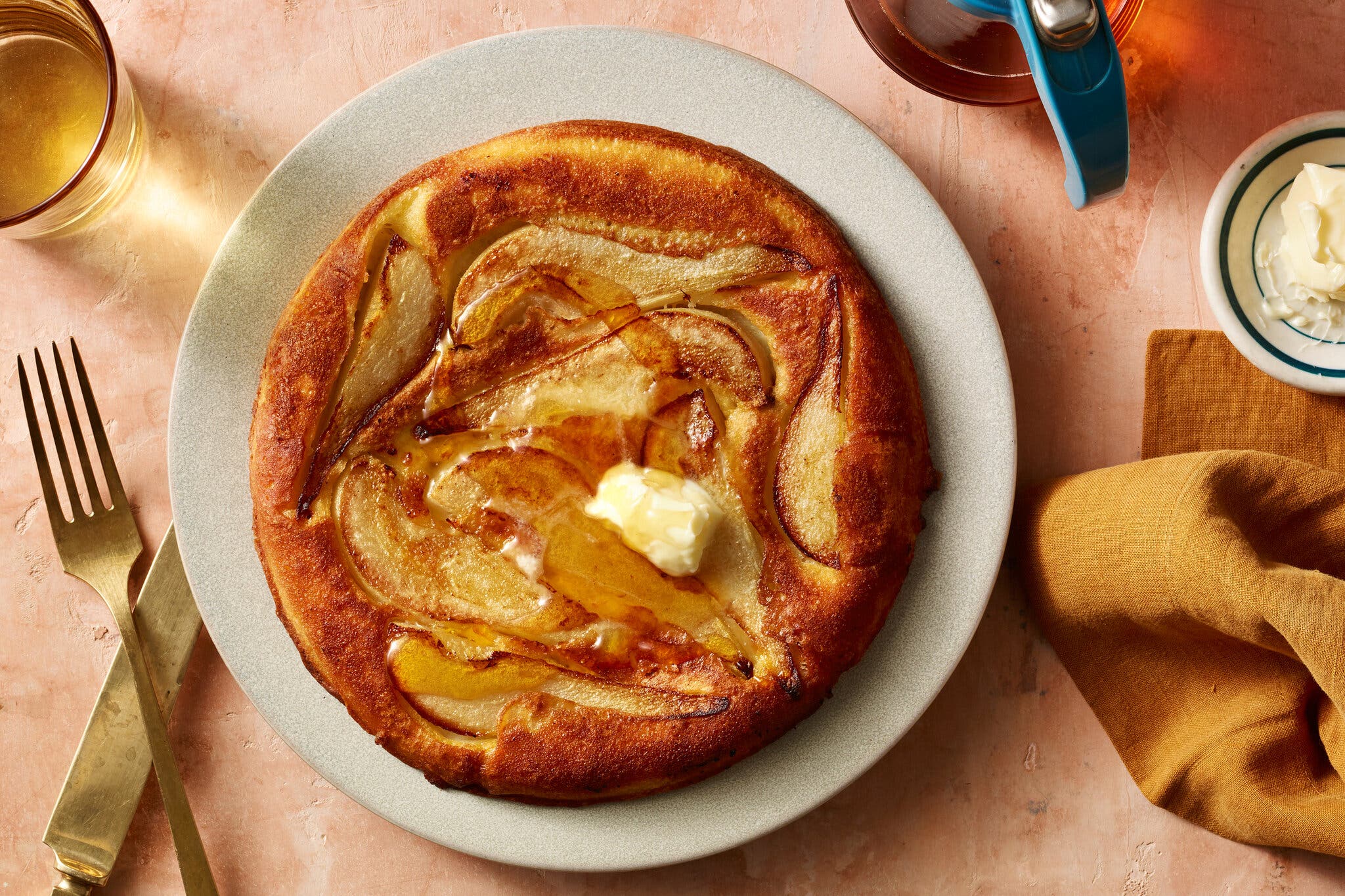 An overhead shot of a large, round pancake topped with sliced pears, a pat of butter and a drizzle of syrup, on a white plate.