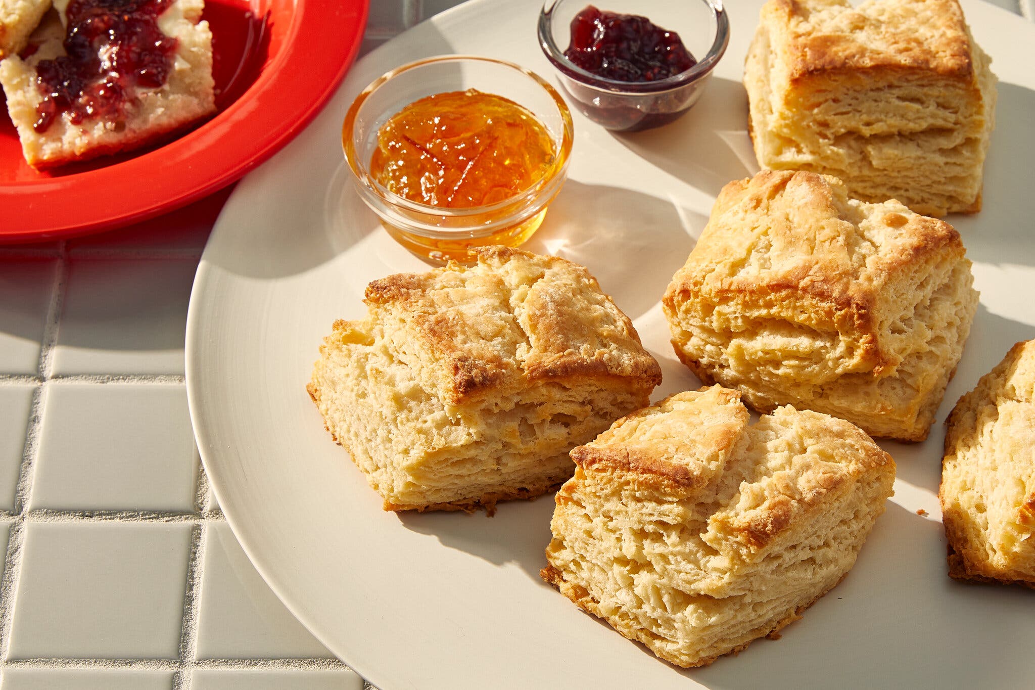 An overhead shot of golden-brown biscuits on a plate with small bowls of orange marmalade and red jam.