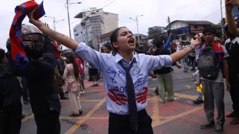 BBC A young woman, wearing what resembles a school uniform which has been emblazoned with protest slogans, shouts and holds her arms and the Nepali flag in the air