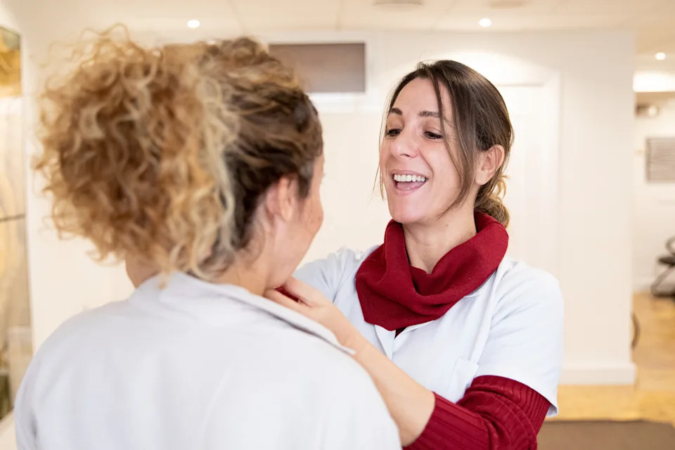 Two women in a workplace setting, one adjusting the other's scarf, both smiling and interacting warmly