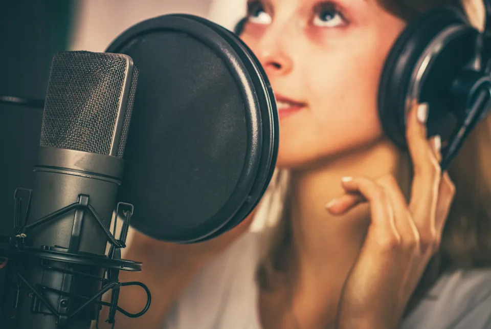 A person listens intently with large headphones in front of a microphone, possibly in a recording studio setting