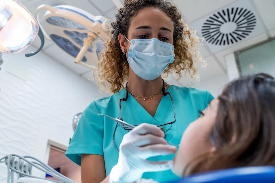 Dentist in scrubs and mask examining a patient's teeth in a clinic setting