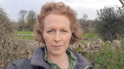 Sarah Godwin Sarah, wearing an outdoor jacket and green shirt, stares down the lens, in a selfie-style picture taken in front of a traditional stone wall, with fields in the distance.