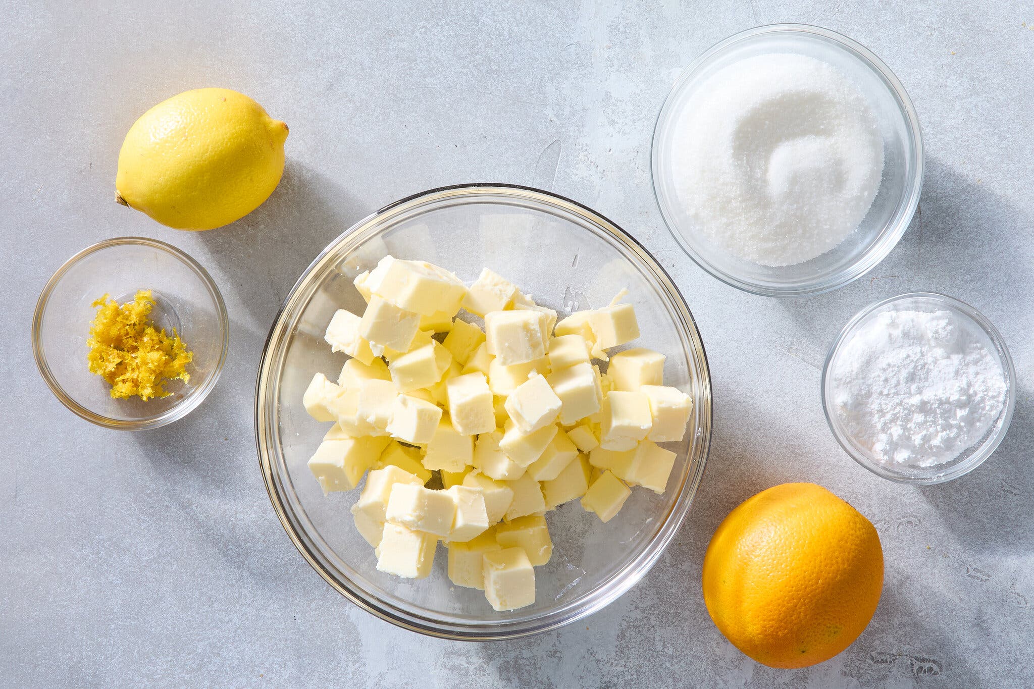 An overhead image of various ingredients in clear bowls, including granulated sugar, lemon zest, cubed butter and powdered sugar. A lemon and an orange sit nearby.