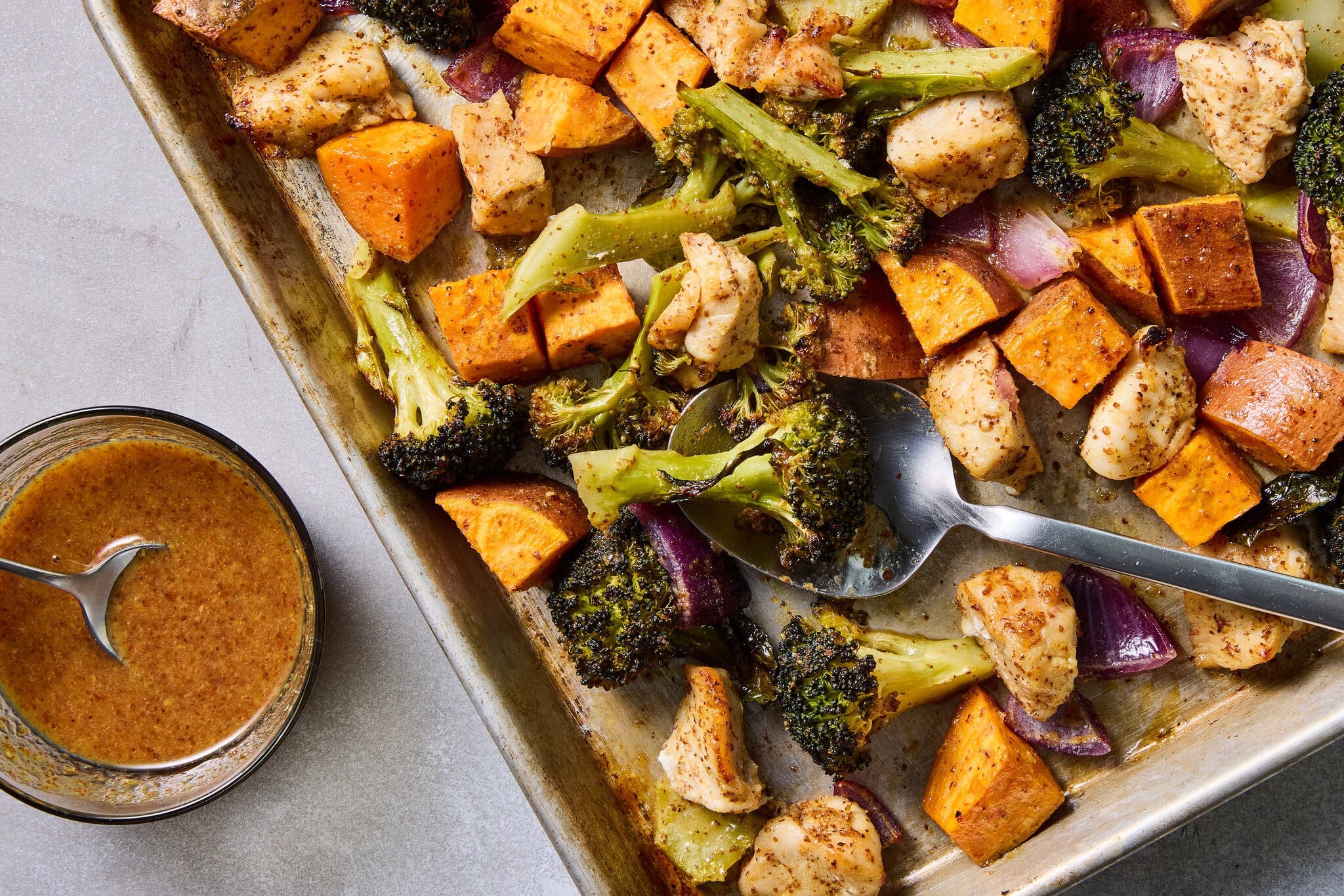 An overhead image of a sheet pan topped with chicken, broccoli and sweet potatoes. A small bowl sits off to the side.