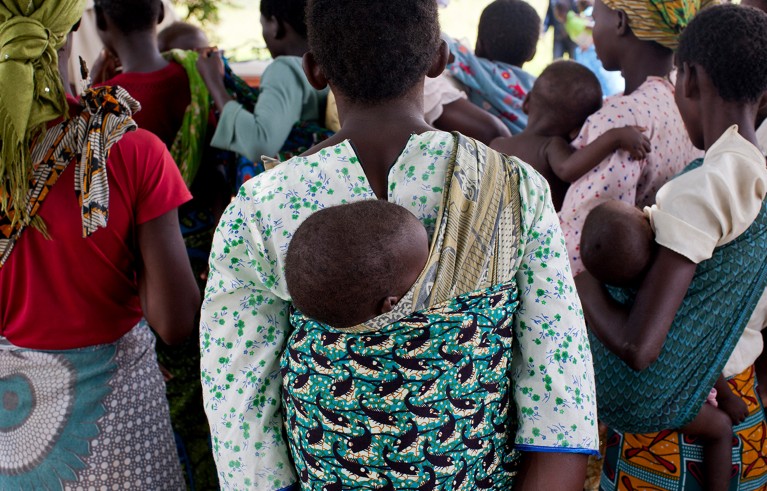 Black mum carrying baby on her back in a sling while waiting in a crowd.